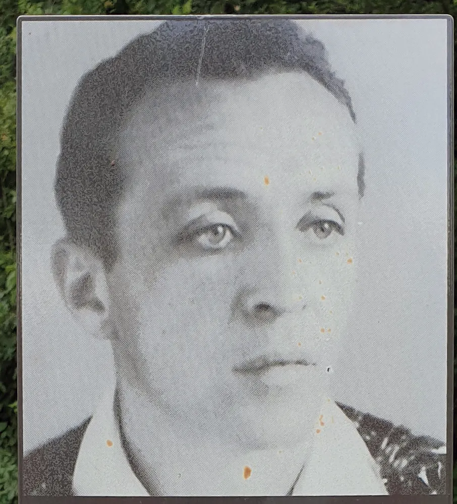 Black-and-white portrait photograph of a man with short dark hair and an open collar, reproduced on a weathered panel