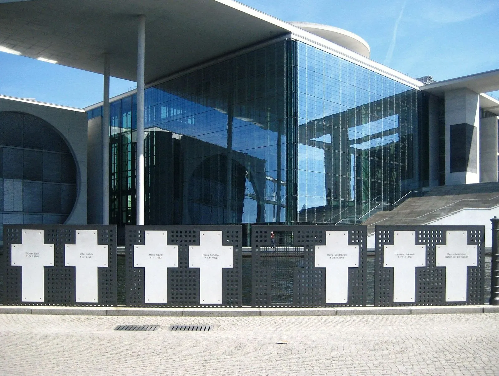 Row of white metal crosses set in dark perforated panels along a riverside railing, in front of a large modern glass building