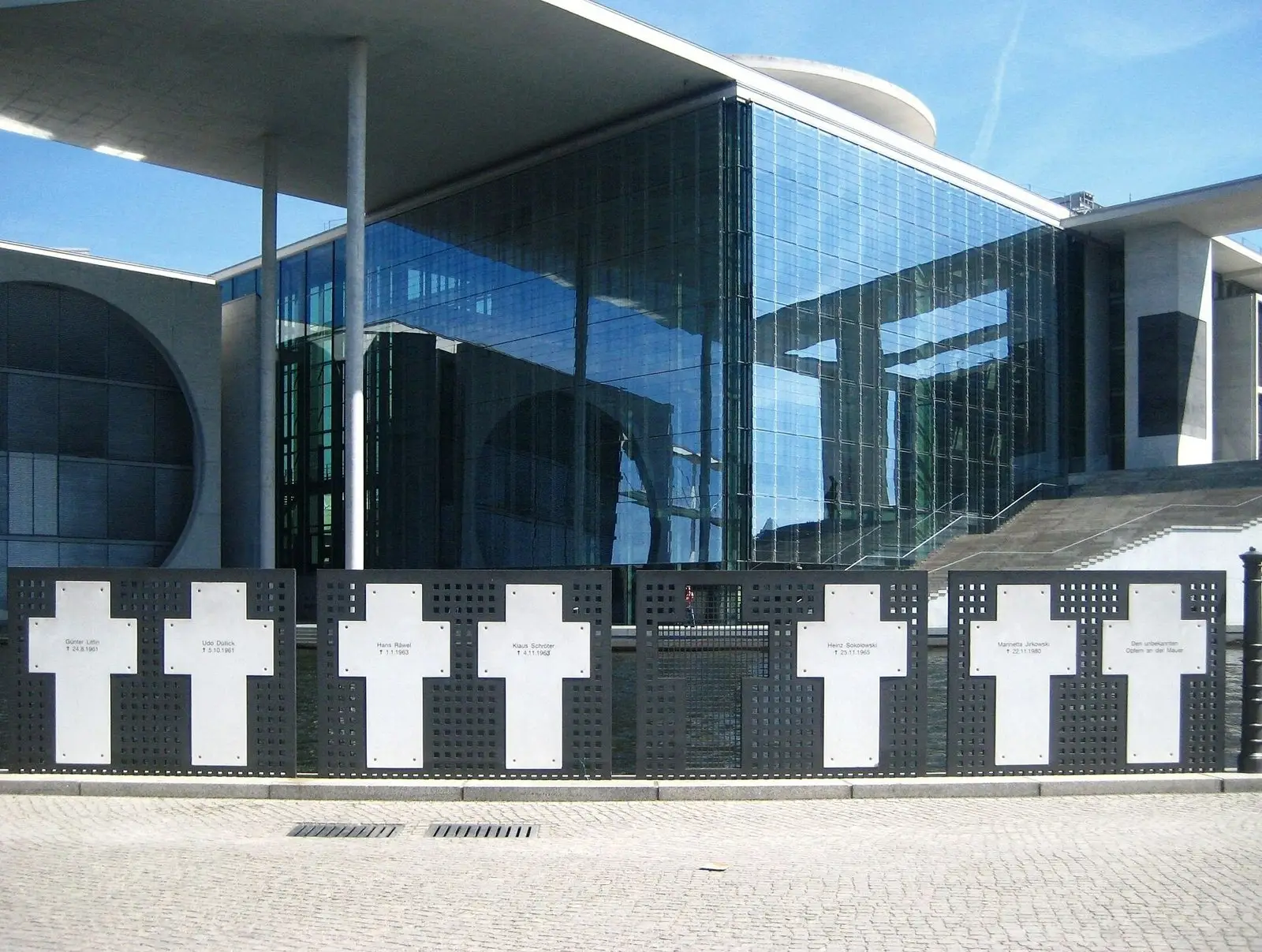 Row of white metal crosses set in dark perforated panels along a riverside railing, in front of a large modern glass building