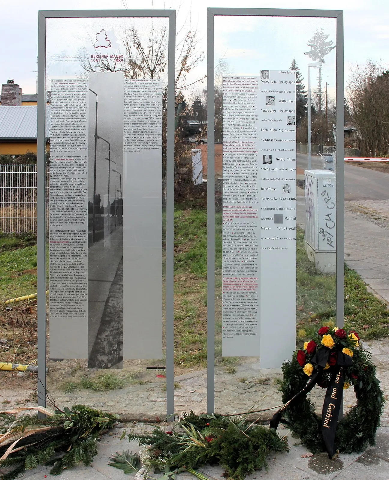 Two glass panels in steel frames with text and small portraits, wreaths laid in front