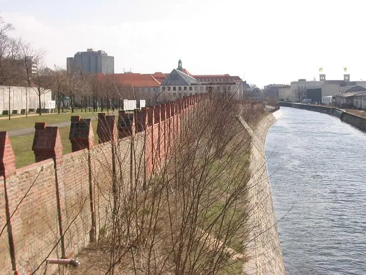 A canal with a sloping stone embankment runs into the distance; on the left a red-brick wall with small pointed pillars, a lawn and large buildings, with bare shrubs along the bank.