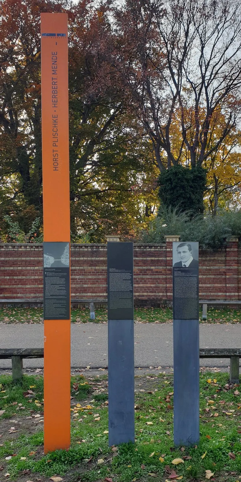 Tall orange column with the names "Horst Plischke · Herbert Mende" and two grey panels in front of a brick wall and autumn trees