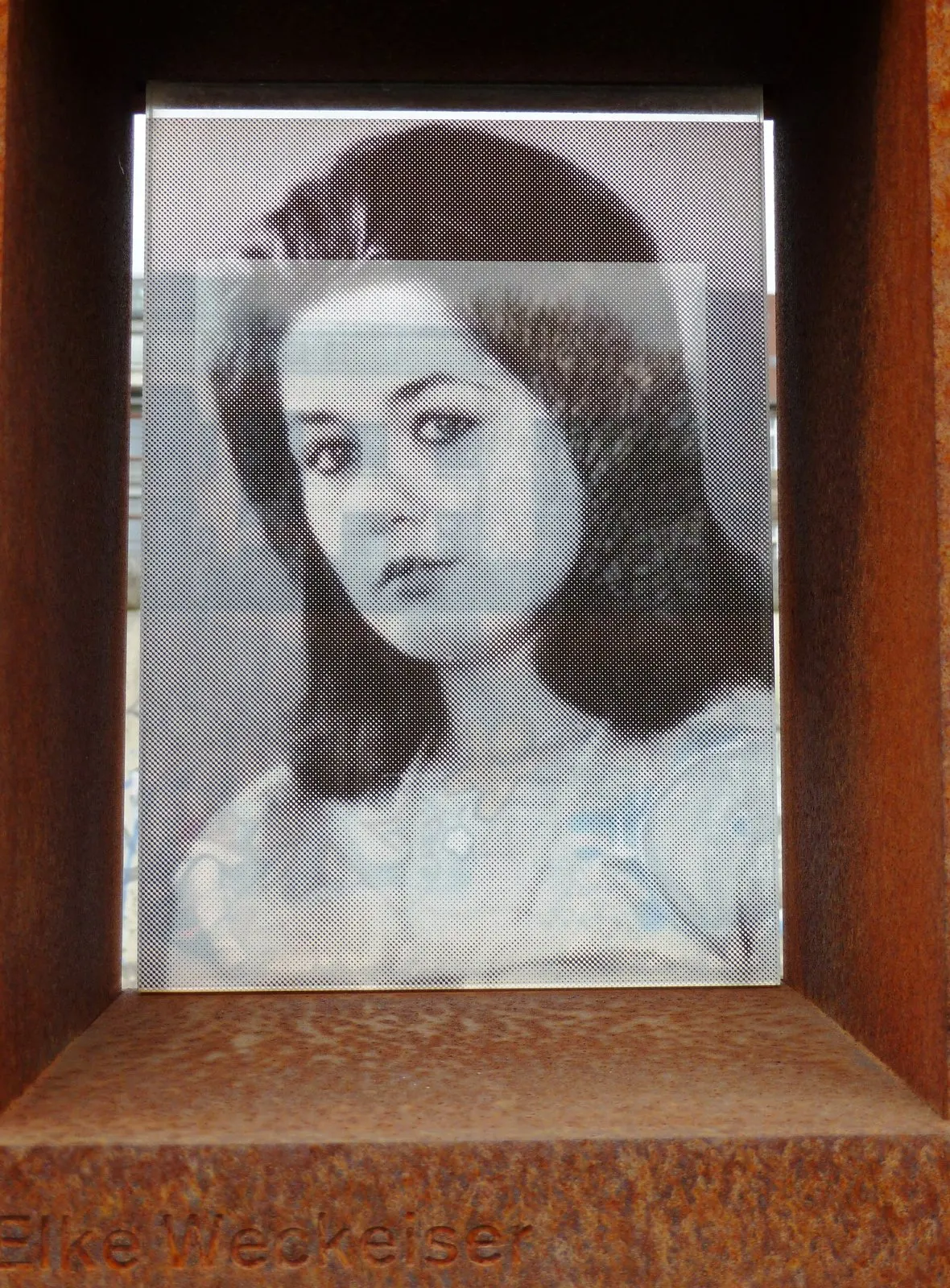 Black-and-white portrait of a young woman with long dark hair, printed on glass inside a rusted steel frame, with the name "Elke Weckeiser" cut into the steel below