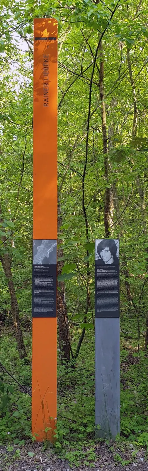 Tall orange column bearing his name in a wood, next to a grey post with a portrait of a young man with long dark hair and a text panel