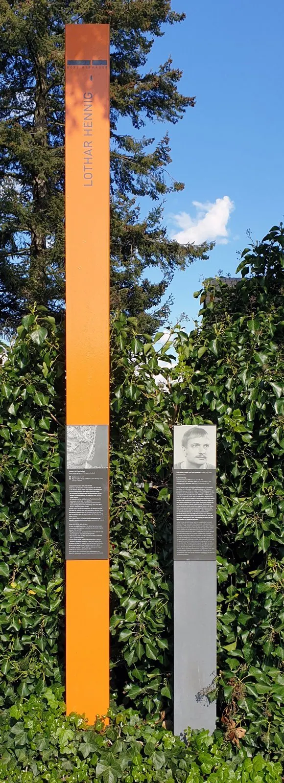 Tall orange column bearing his name in front of an ivy hedge, next to a grey post with a portrait and text panel