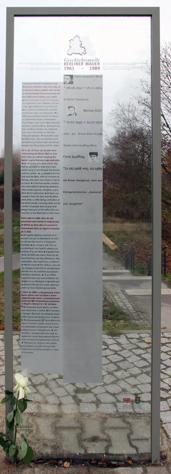 Tall glass panel in a metal frame printed with text and three small portraits with names and dates, a white rose at its foot, beside a paved path