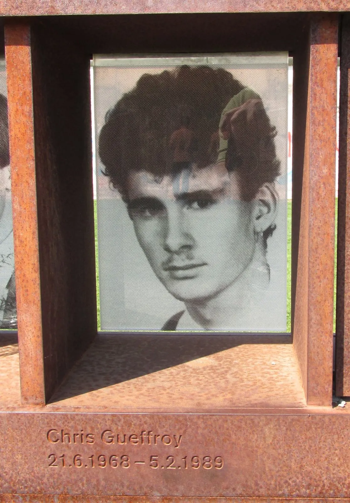 Black-and-white photograph of a young man with thick dark hair, set in a rusted steel niche engraved with his name and dates