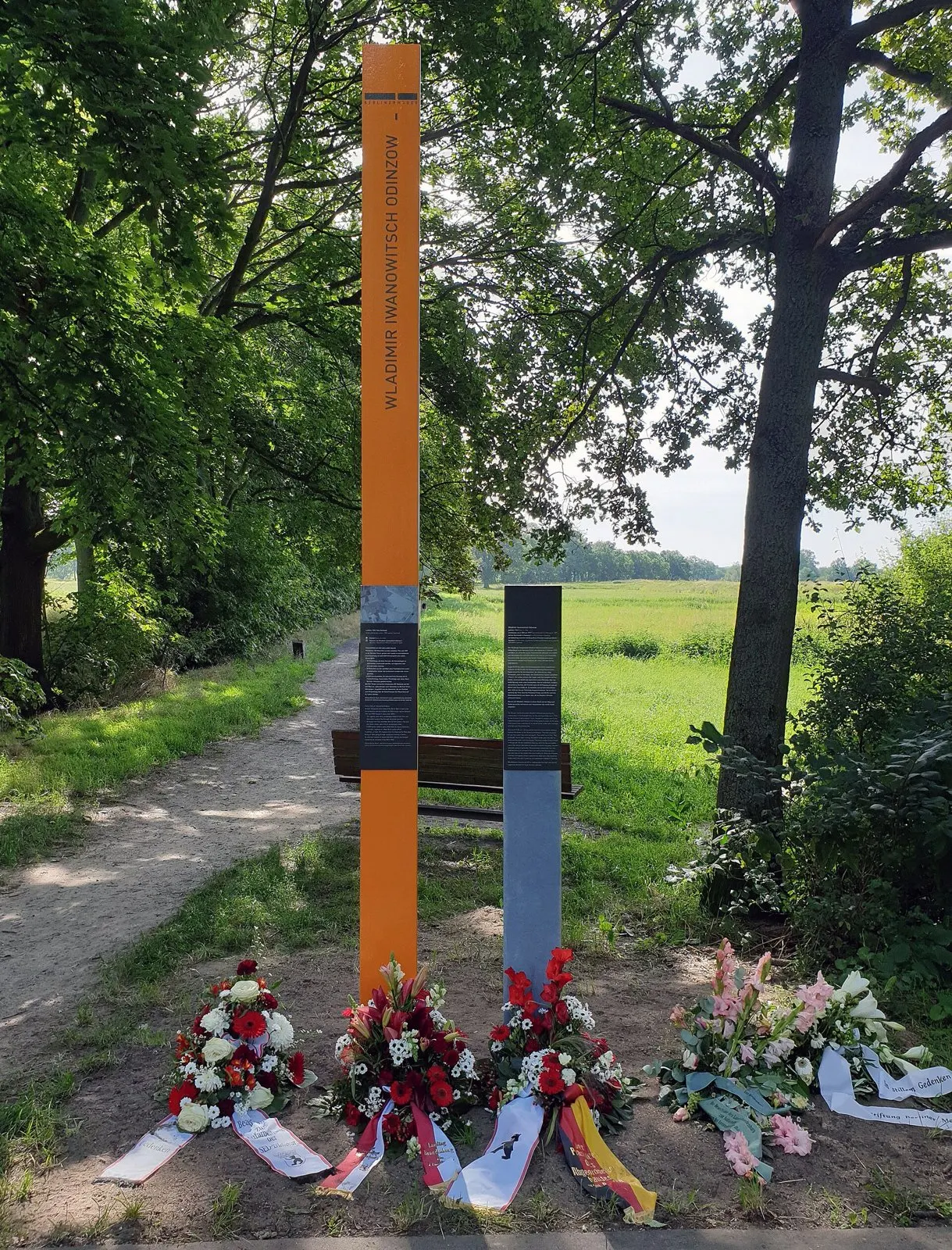 Tall orange column with the name Wladimir Iwanowitsch Odinzow and a grey text post beside a path at the edge of a field, with wreaths and flowers at the foot