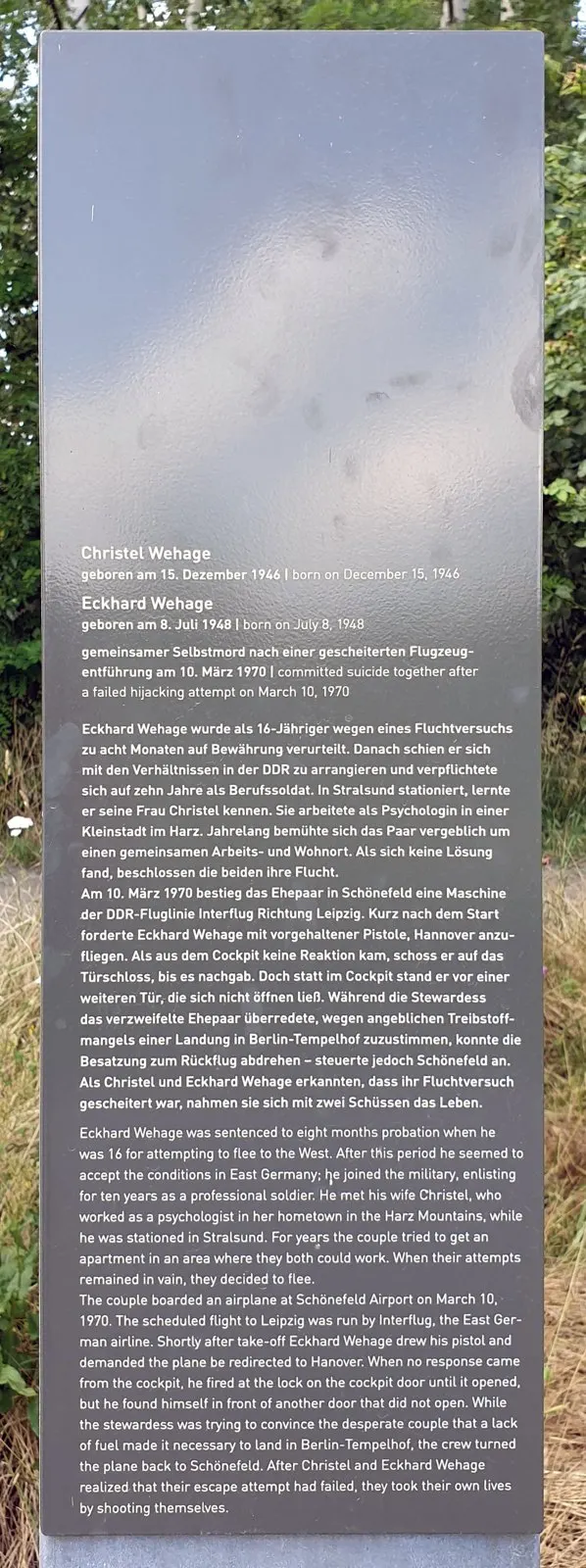 Dark stele with printed German and English text headed by the names Christel Wehage and Eckhard Wehage, in front of bushes and trees