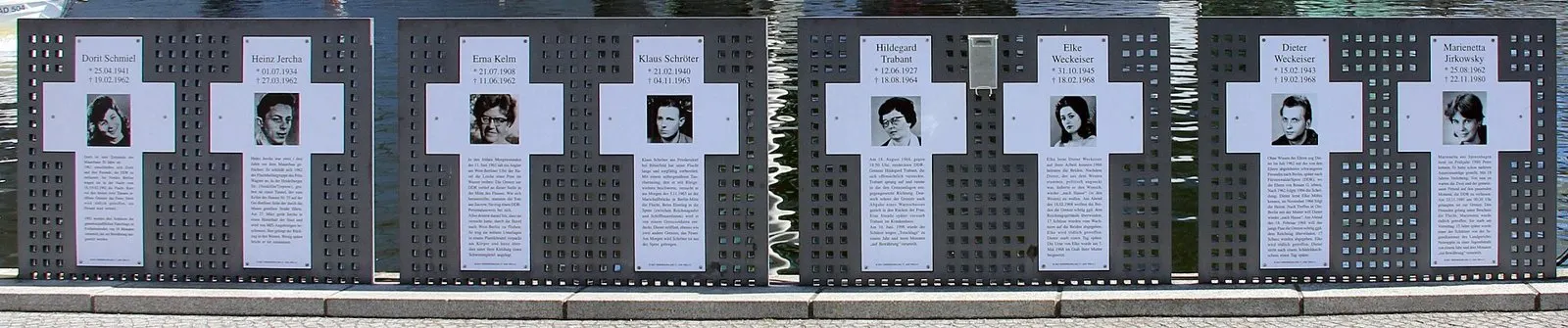 Row of white crosses with small portrait photographs, names and text, mounted on perforated dark metal panels in front of water