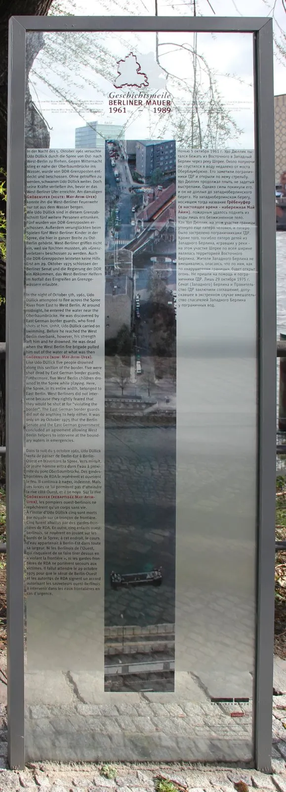 Tall glass panel in a steel frame with text in four languages around a vertical strip photograph of the riverbank