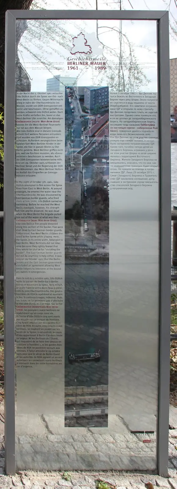 Tall glass panel in a steel frame with text in four languages around a vertical strip photograph of the riverbank