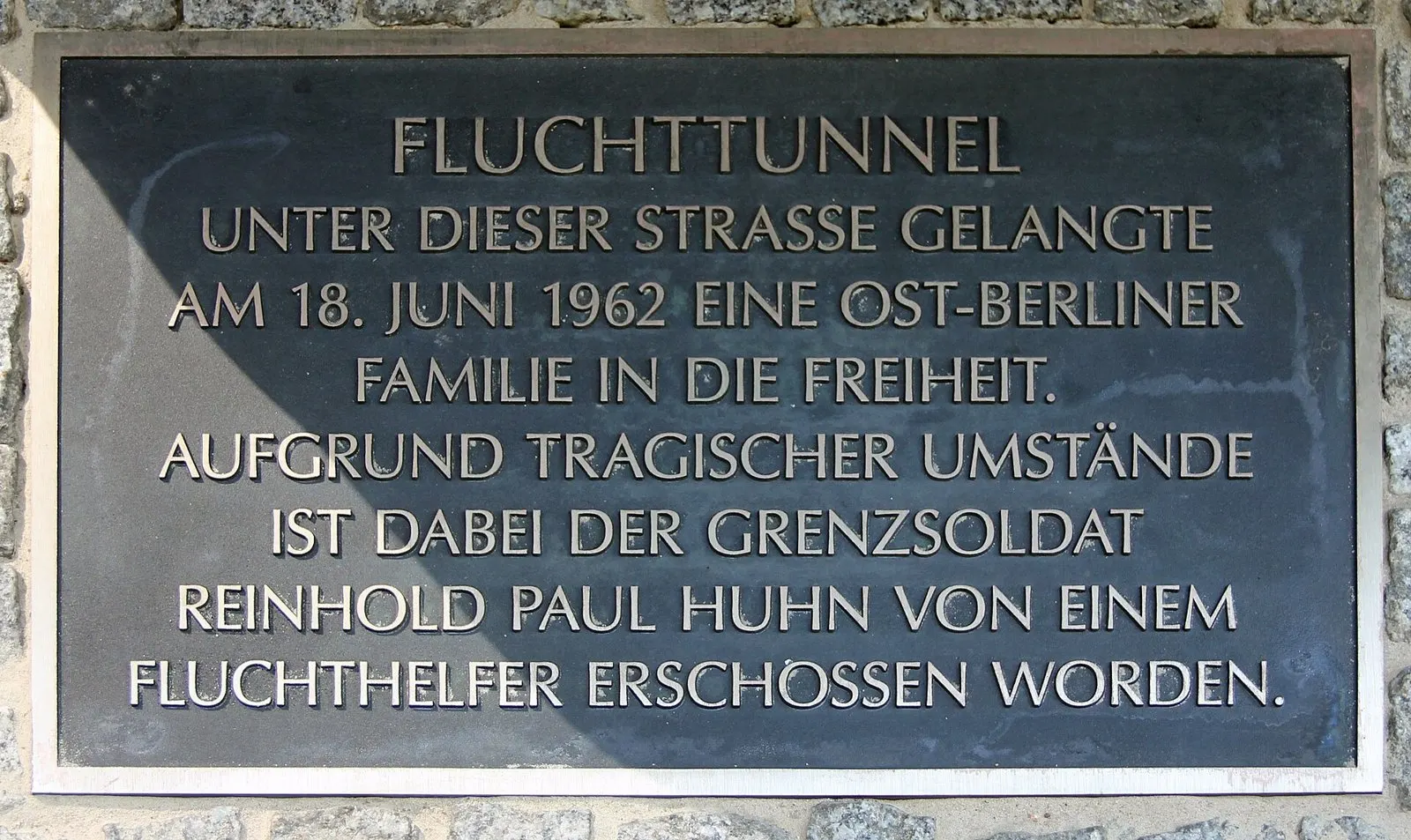 Dark plaque with raised letters stating that an East Berlin family escaped through a tunnel on 18 June 1962 and that the border soldier Reinhold Paul Huhn was shot