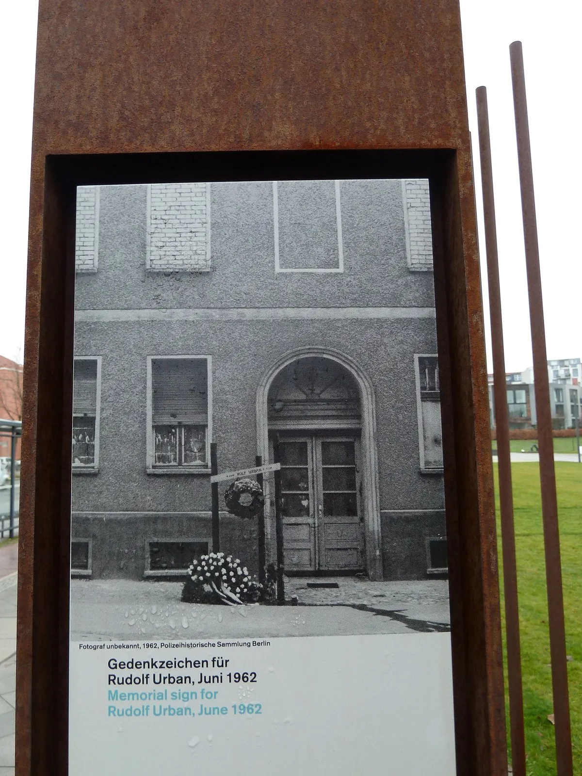 Rusted steel stele framing a black-and-white photo of a house doorway with a cross and wreaths, captioned "Gedenkzeichen für Rudolf Urban, Juni 1962"