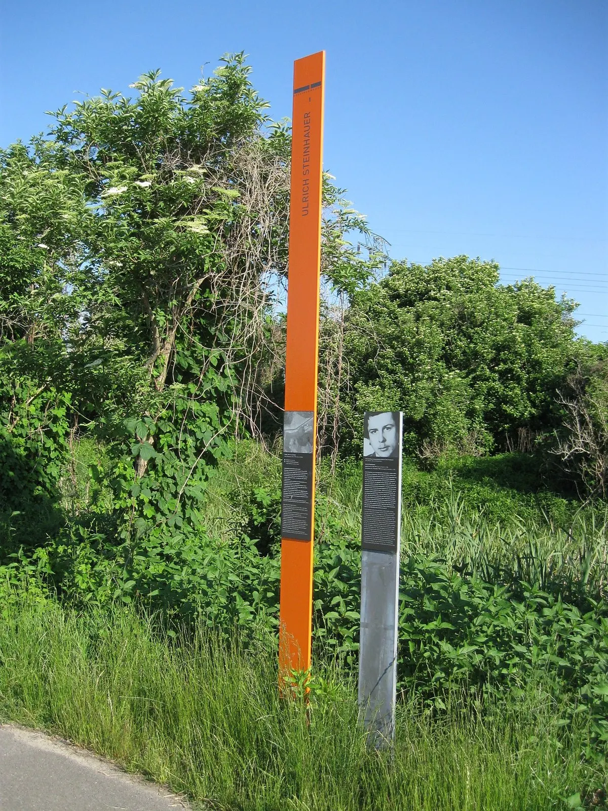 Tall orange column with his name beside a paved path in green scrub, next to a grey post with a portrait panel