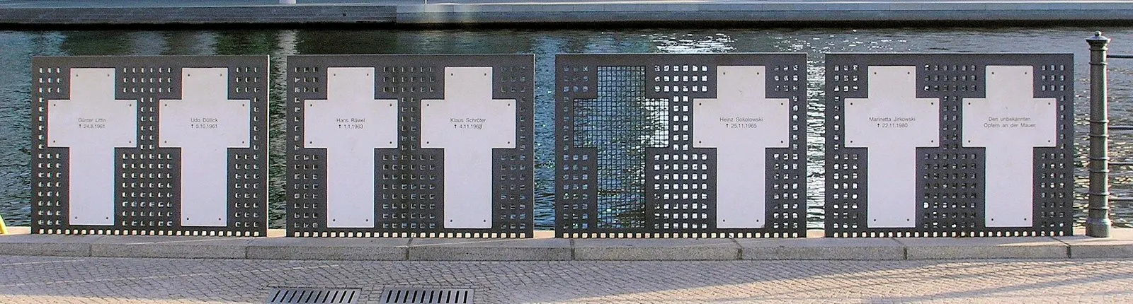 Several white crosses on dark perforated panels in a row along the riverbank