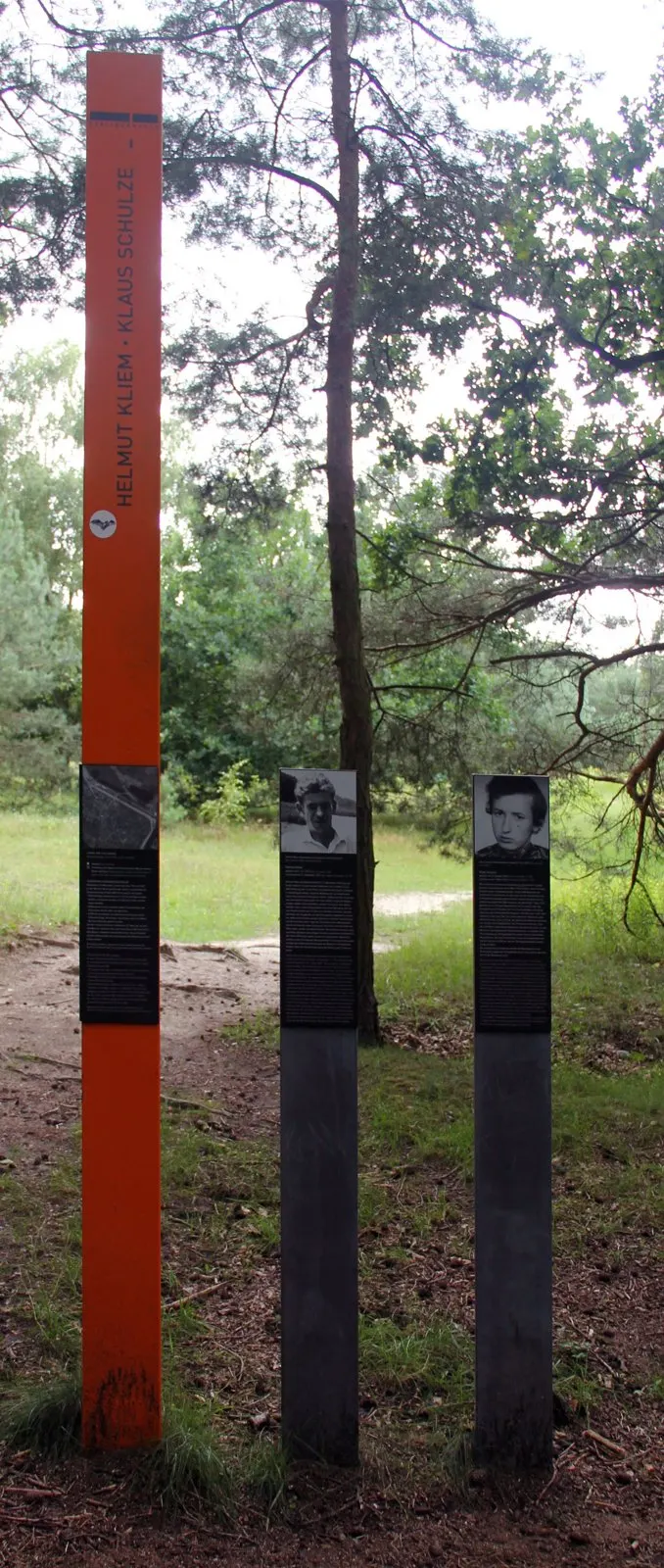 Tall orange column bearing the names Helmut Kliem and Klaus Schulze beside two dark steles with portraits, on a forest path