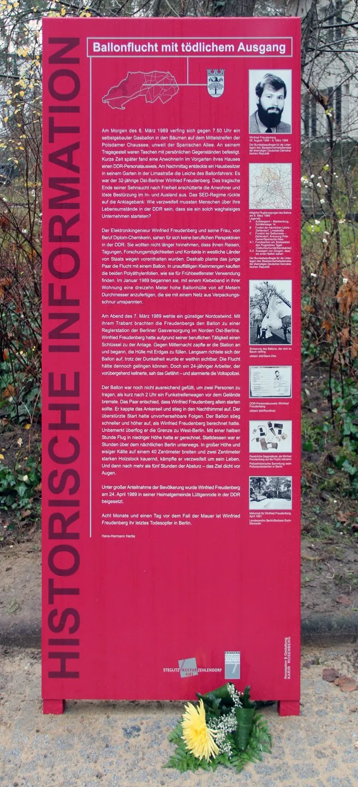 Tall red information panel headed "Historische Information" with text, a map and small photographs, with a yellow flower at its foot