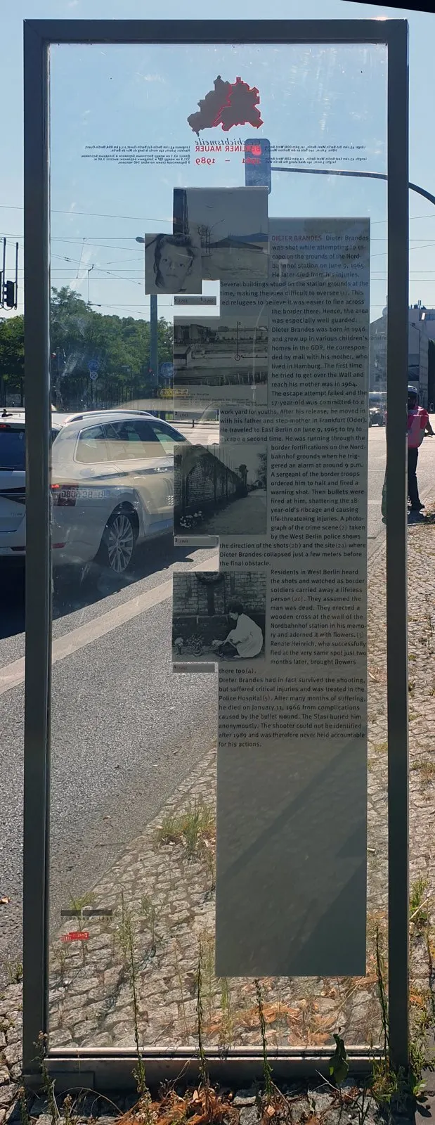Glass panel in a steel frame with English text and small black-and-white photographs, with a street and parked car visible through the glass