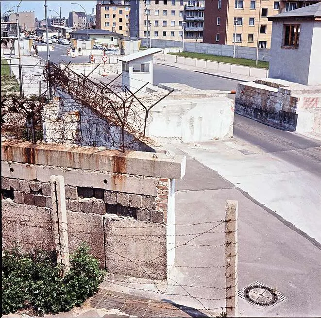 Faded colour photograph looking down on a wall of concrete blocks topped with barbed wire; behind it a checkpoint road with barriers, a small guard hut and apartment blocks.