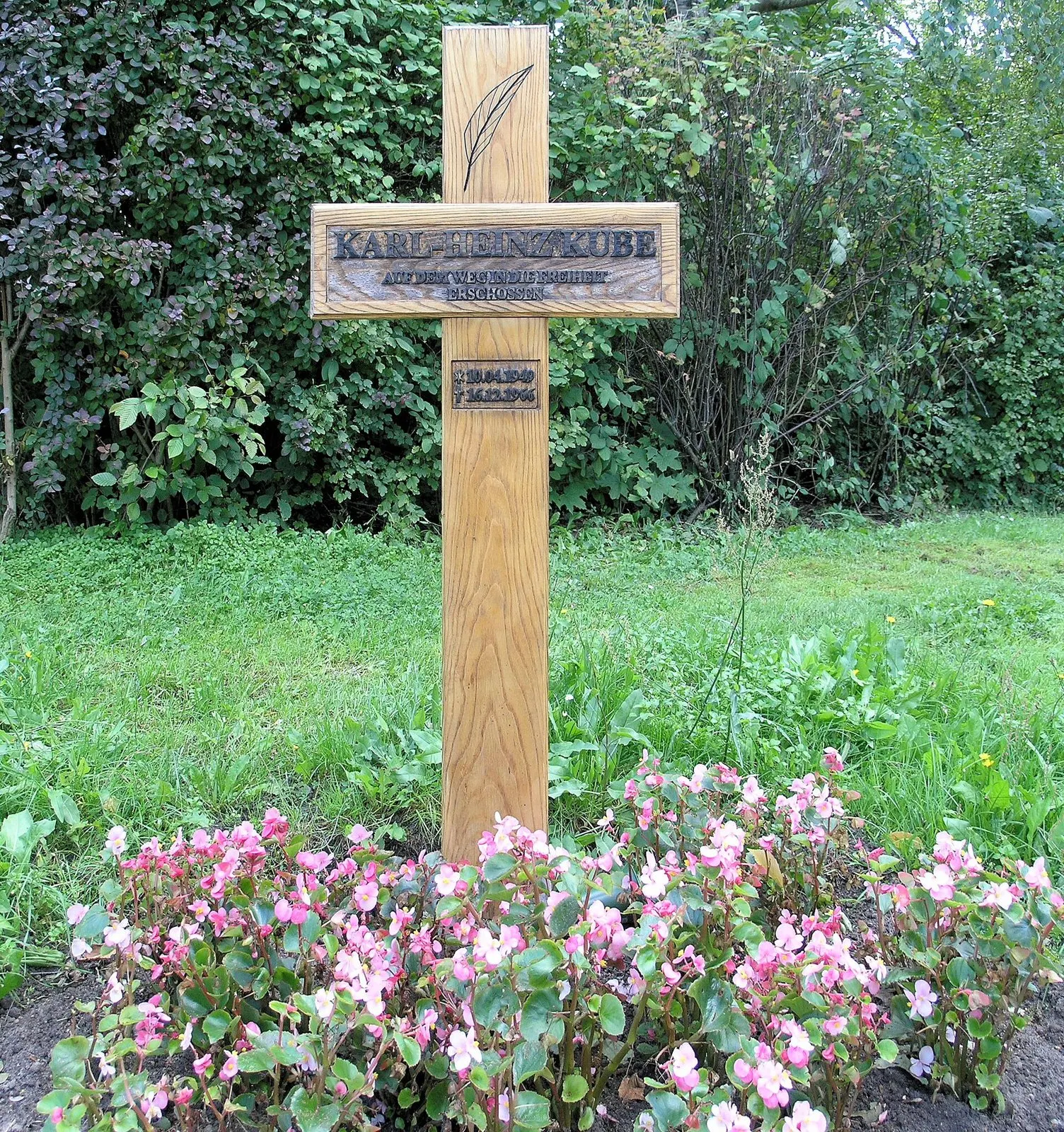 Light wooden cross with a carved name plate "Karl-Heinz Kube" standing in grass above pink flowers