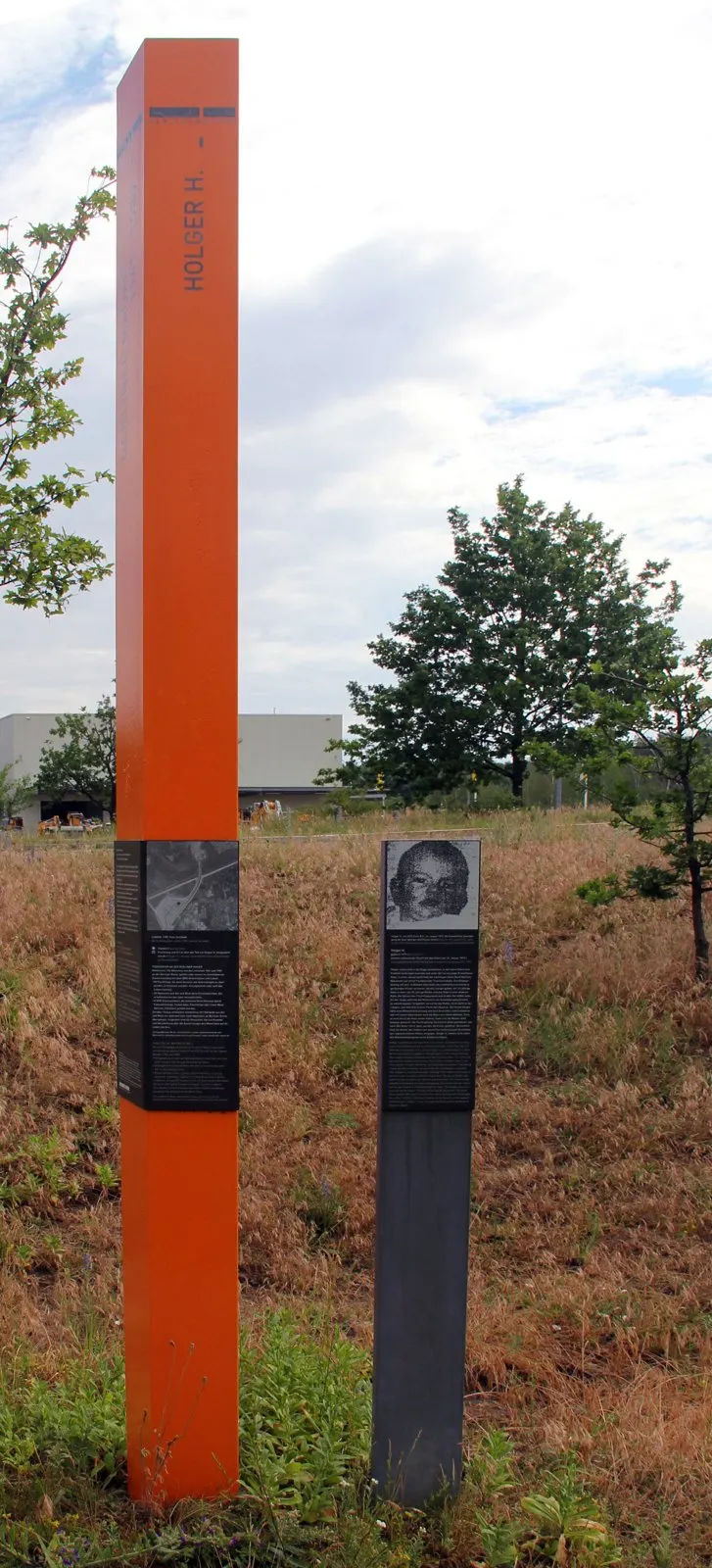 Tall orange column bearing the name Holger H. and a dark stele with a child's portrait, in a field of dry grass