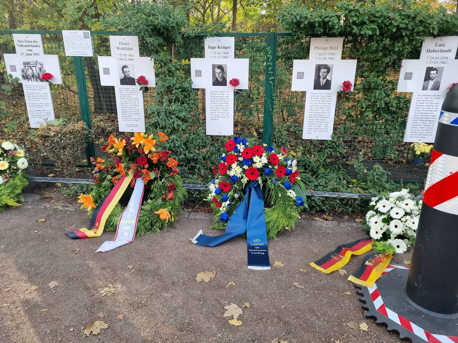 White crosses on a green fence with flower wreaths in front; the central cross bears the name Ingo Krüger, a black-and-white portrait of a young man and a text about his escape attempt through the Spree in diving gear