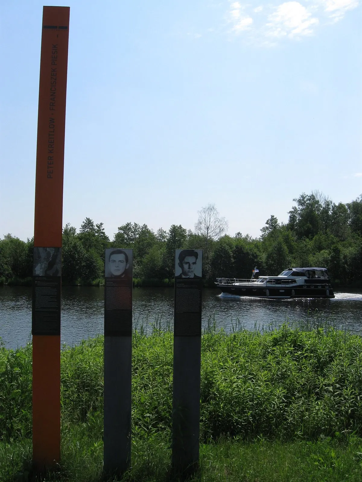 Orange column with two names and two grey panels with portraits on a grassy riverbank, a motor boat passing on the river