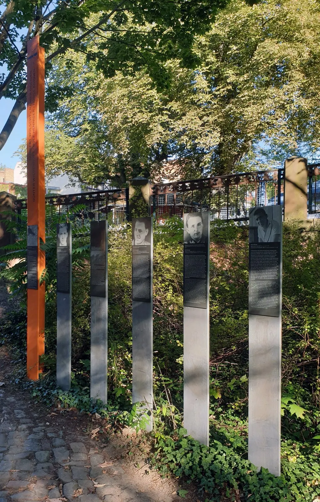Tall orange column and five grey panels, some with portrait photographs, beside a cobbled path under trees