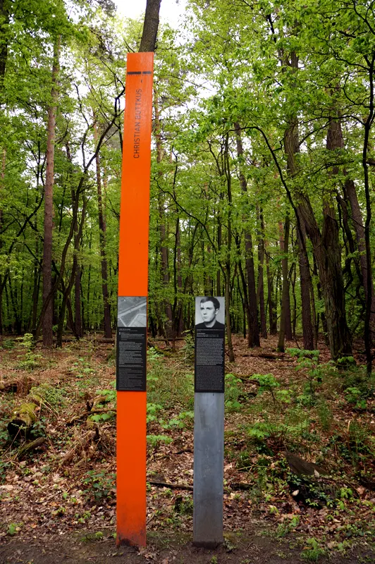 Tall orange column inscribed 'Christian Buttkus' and a grey post with his portrait panel, in green beech woodland