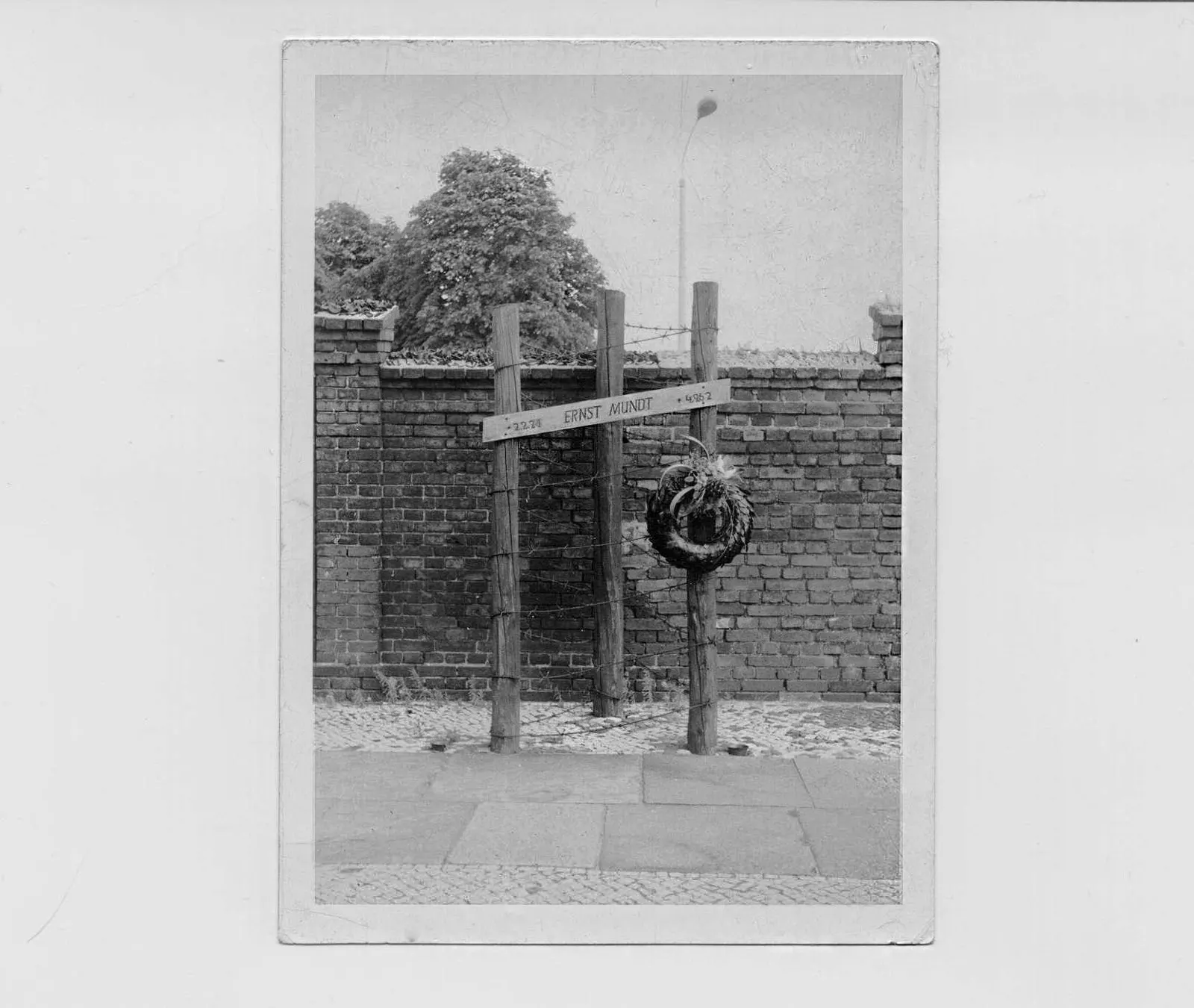 Old black-and-white print of a wooden cross with a wreath and a name board reading "Ernst Mundt" in front of a brick wall topped with barbed wire