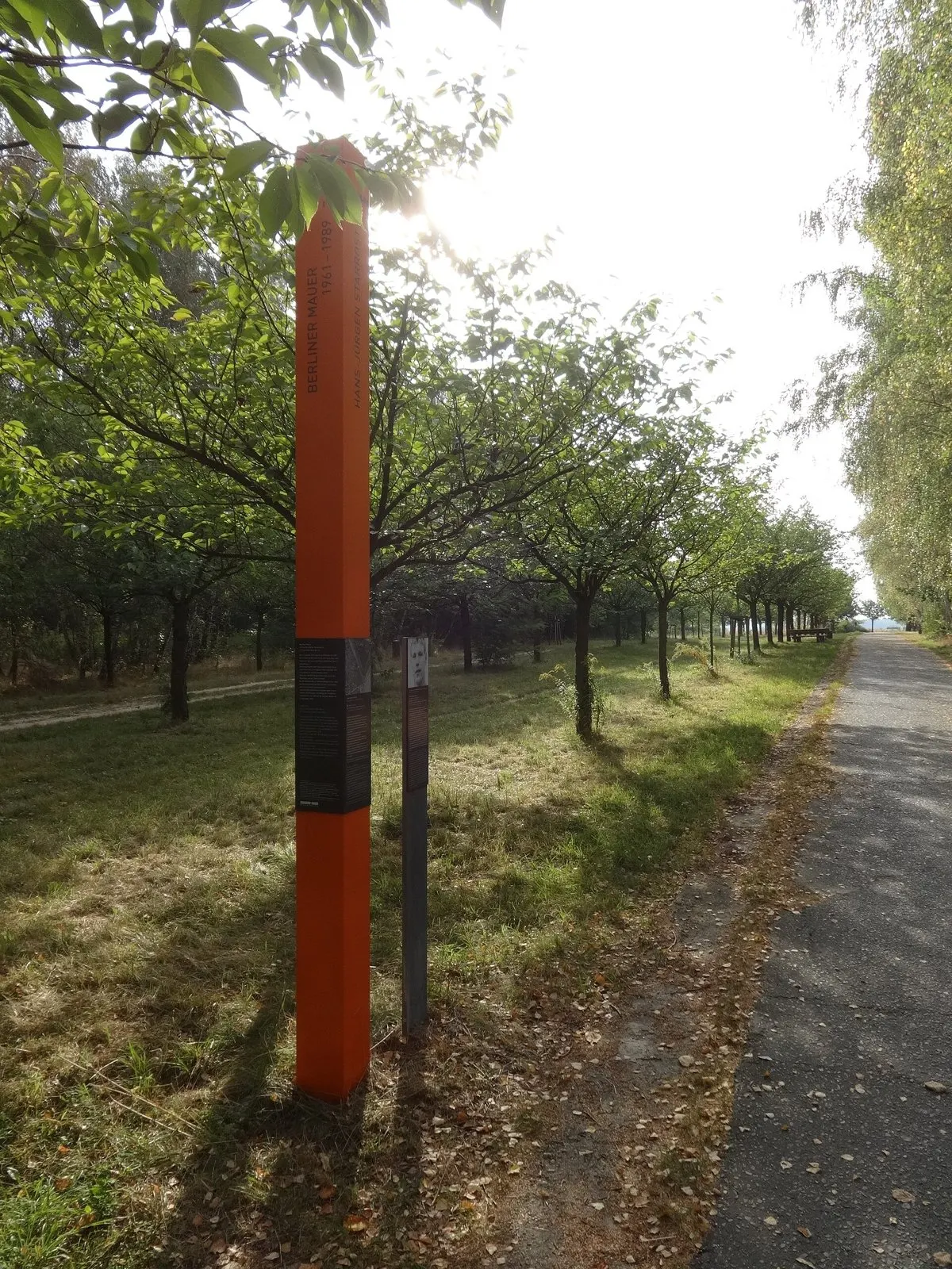 Tall orange column and a slim grey post on grass beside a paved trail lined with young trees