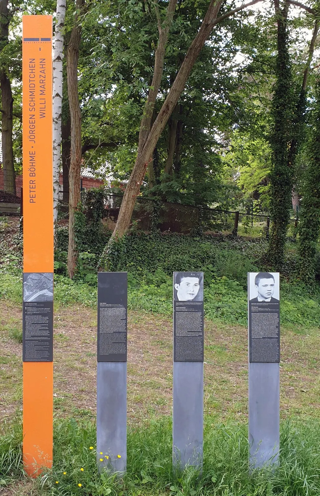 Tall orange column with three names beside three grey panels, two of them with portrait photographs, on a grassy slope under trees