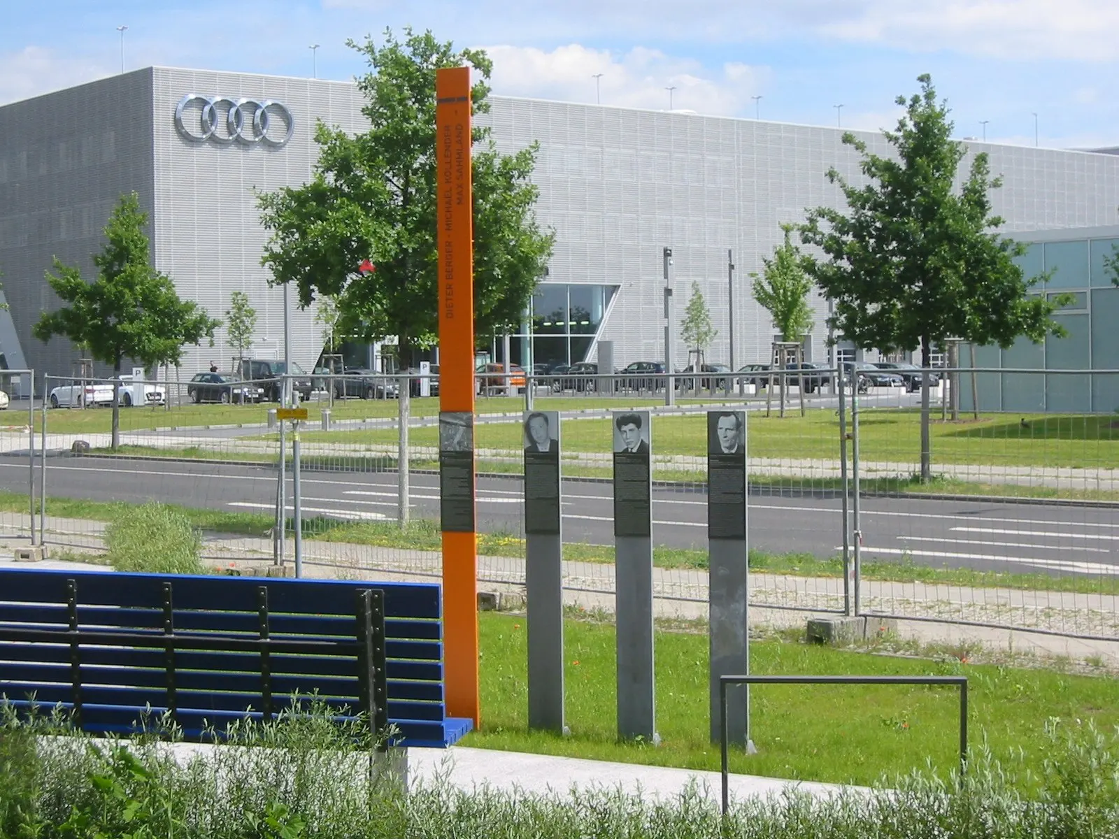 Orange column and three grey steles with portrait photographs on a lawn beside a road, with a bench in front and a large modern building behind