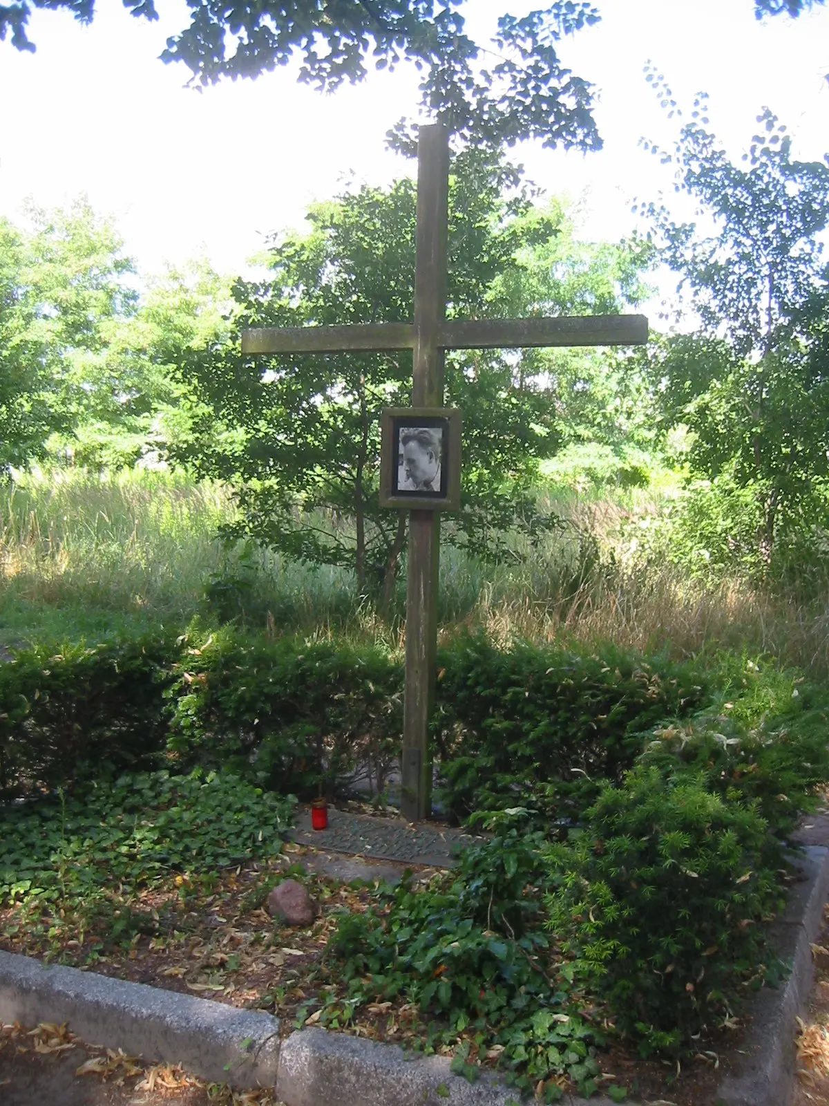 Tall wooden cross with a small framed portrait, surrounded by hedges and ivy