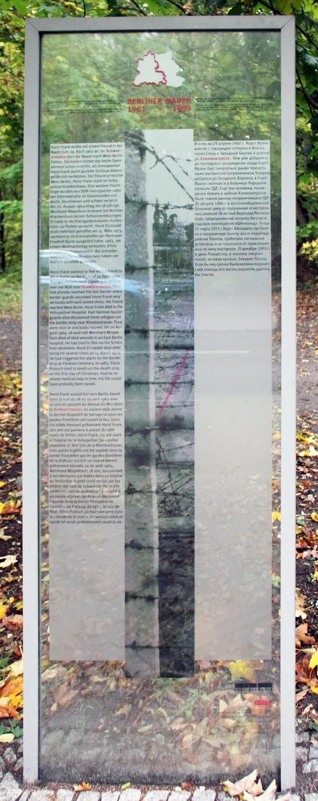 Tall glass panel with multilingual text columns and a barbed-wire photograph, autumn leaves on the ground