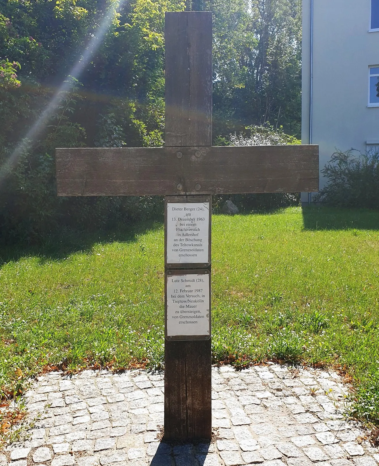 Plain wooden cross on a small cobbled circle on a lawn, with two white plaques bearing text