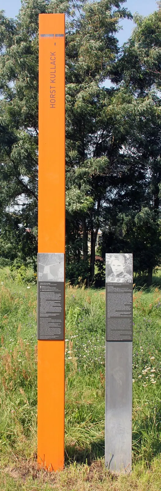 Tall orange column bearing the name Horst Kullack and a dark stele with a portrait, in a meadow with trees