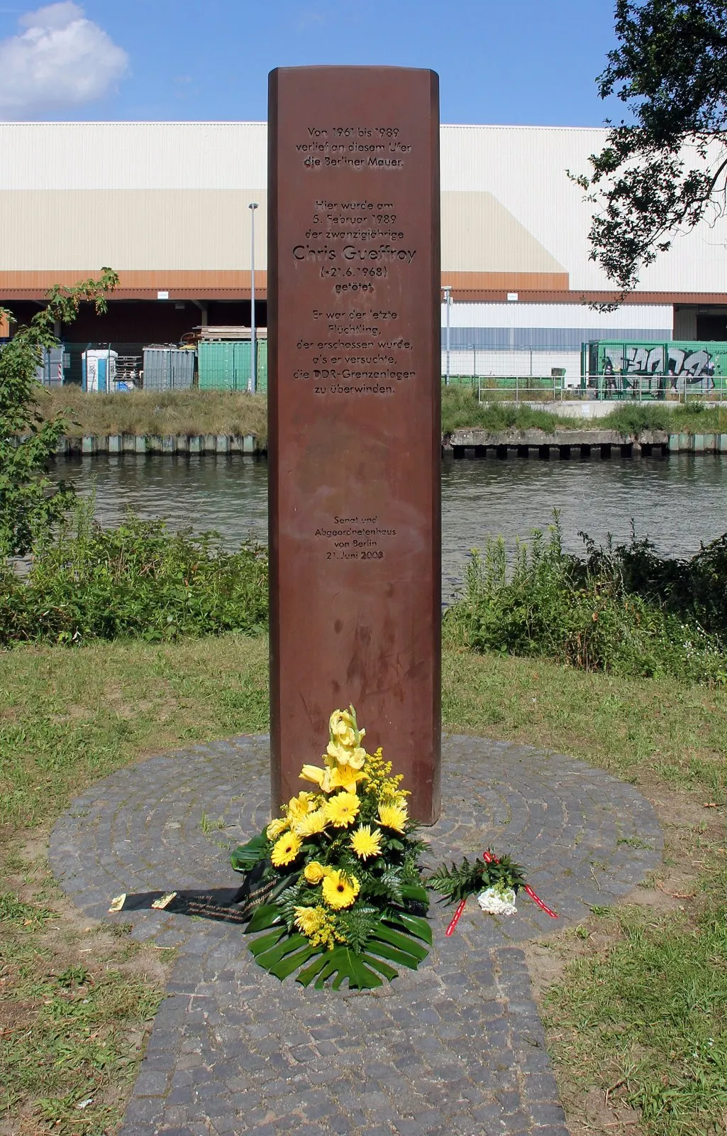 Tall rust-brown steel stele with an engraved inscription on a round paved base with yellow flowers, beside a canal