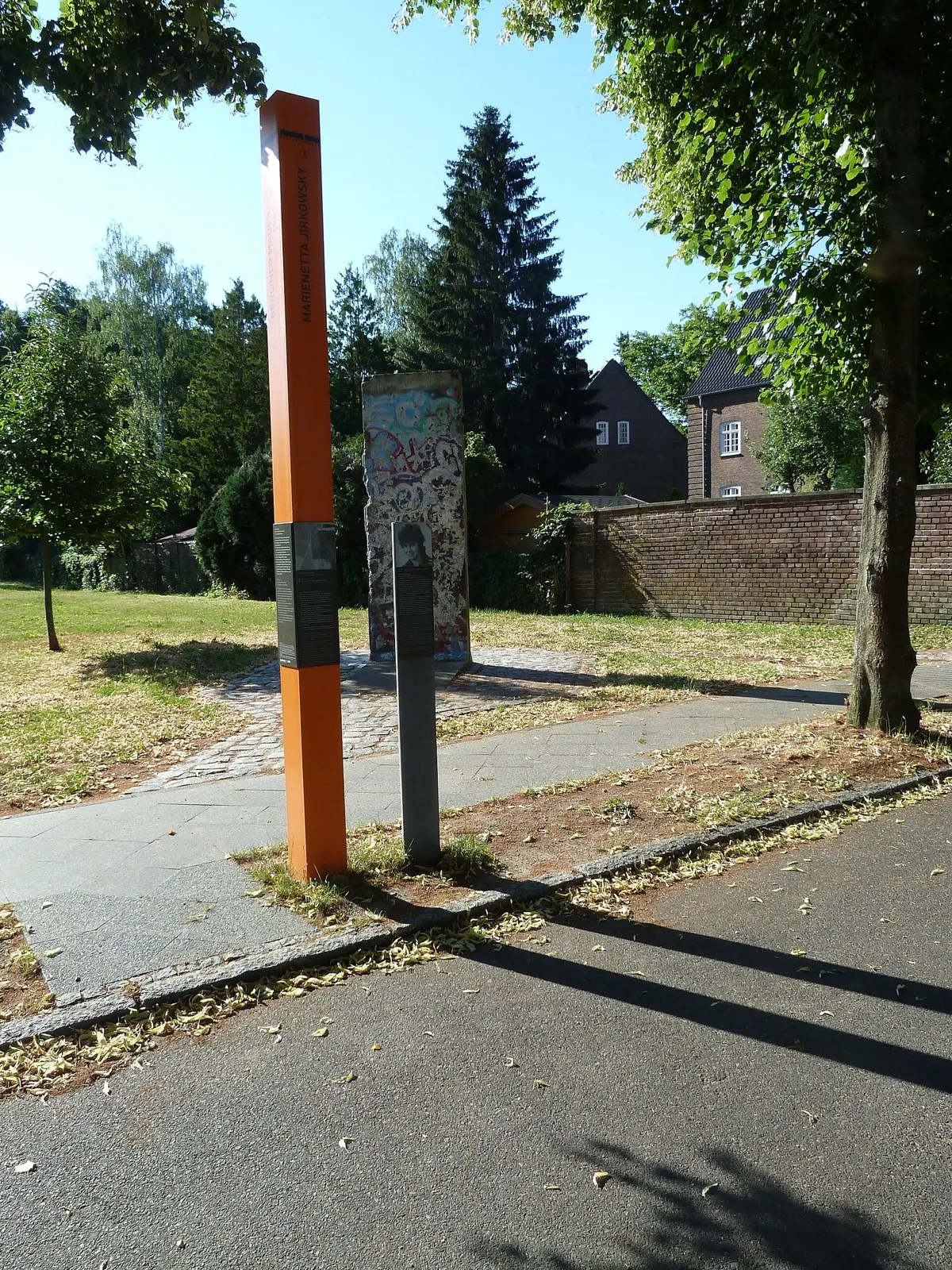 Tall orange column and a grey portrait post beside a path, with a graffiti-covered concrete Wall segment behind them