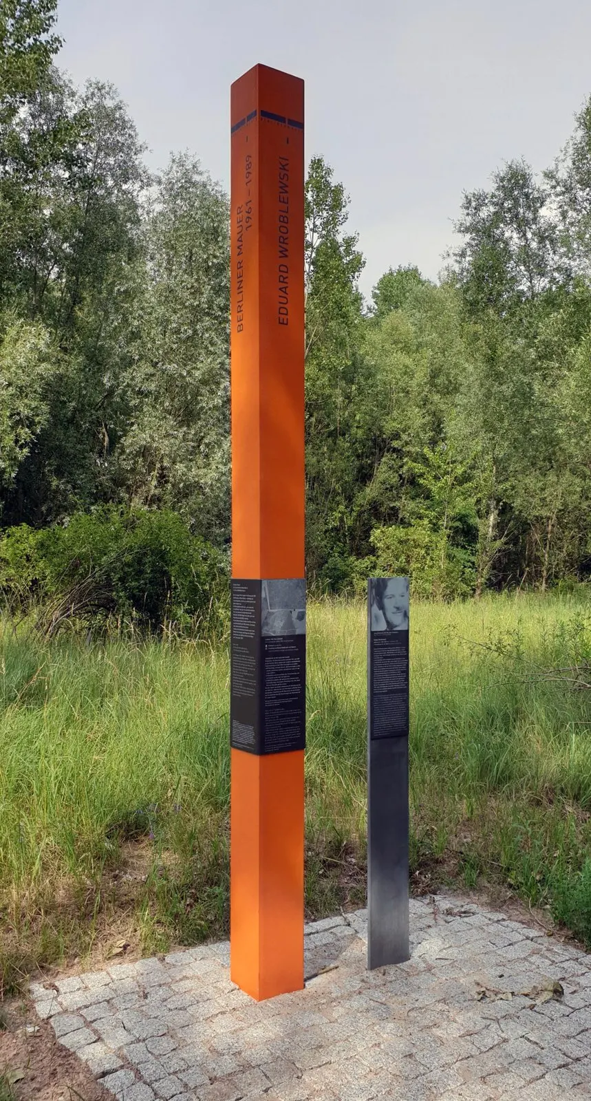Tall orange column with the name "Eduard Wroblewski" beside a grey panel with a portrait, on paving at the edge of a meadow with trees