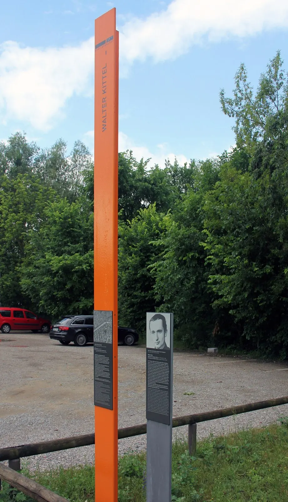 Tall orange column with the name "Walter Kittel" and a grey panel with a portrait photograph, beside a wooden fence and a gravel car park