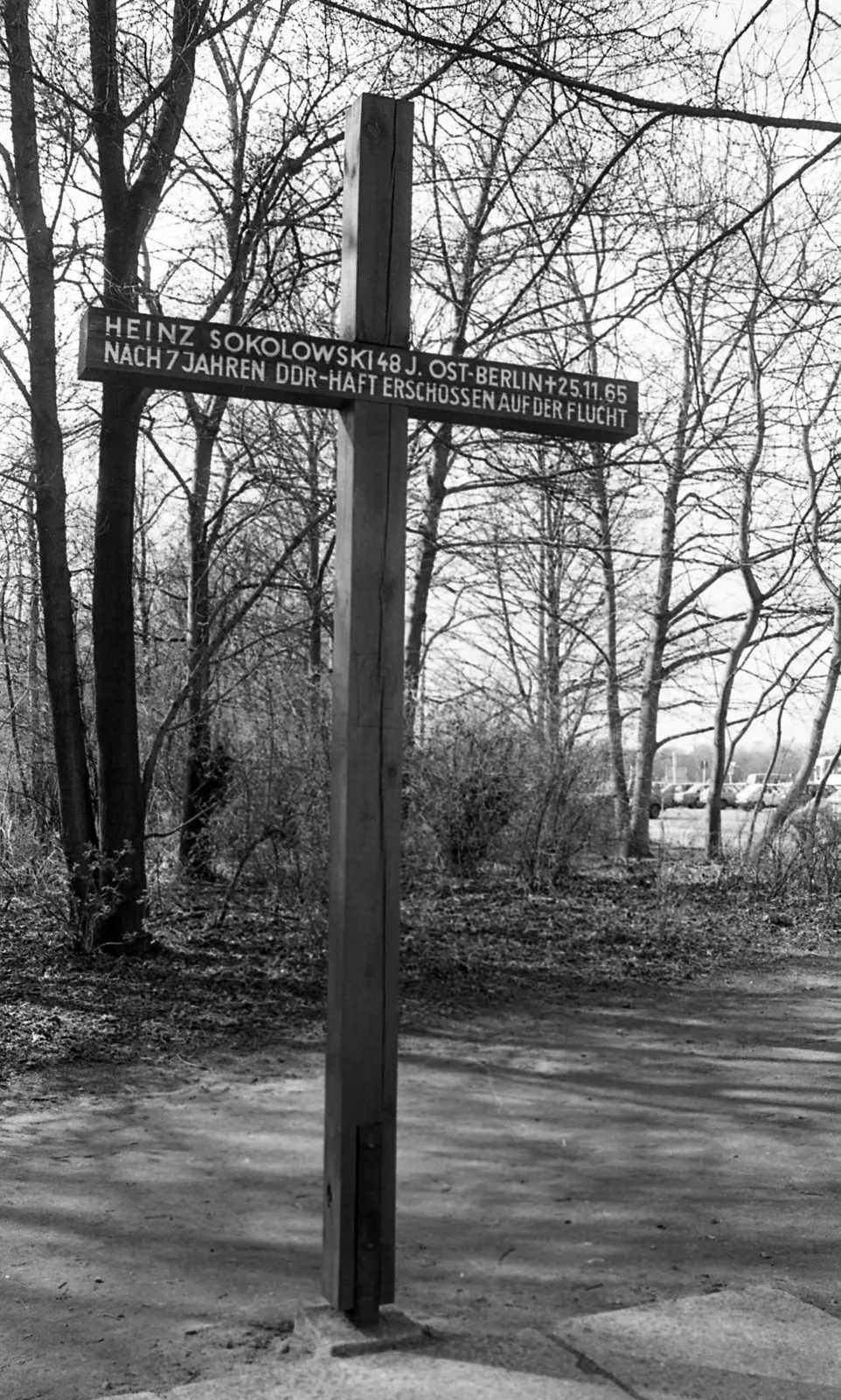 Black-and-white photograph of a tall wooden cross with a sign bearing the name "Heinz Sokolowski", among bare trees
