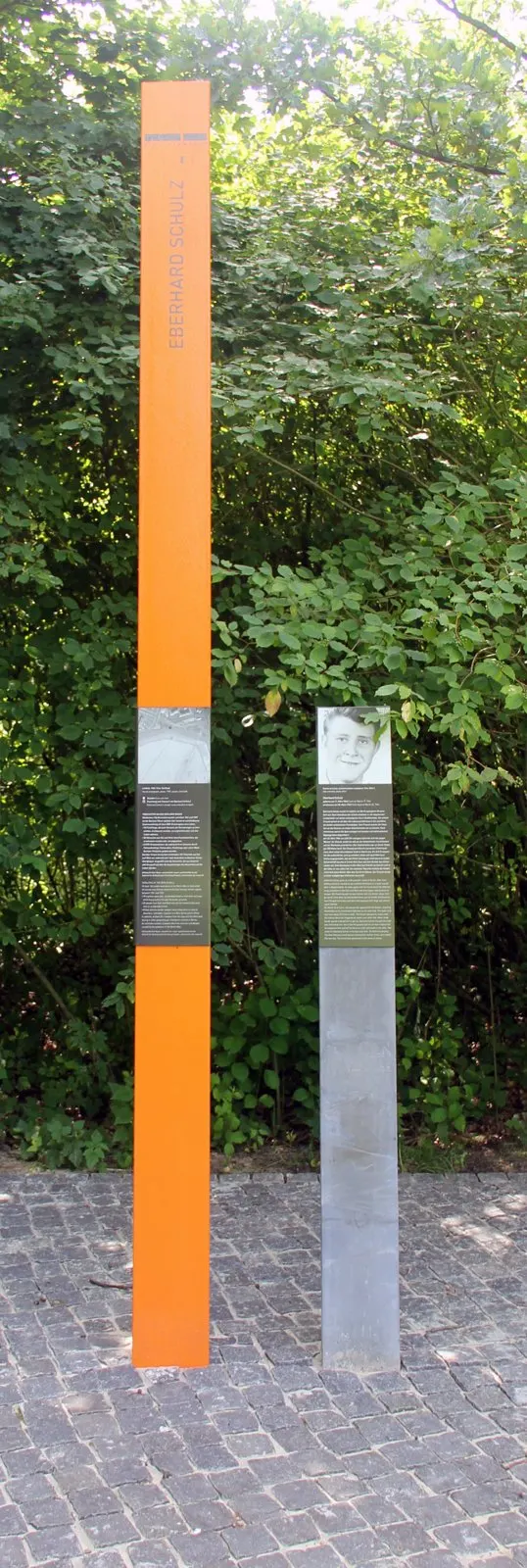 Tall orange column bearing the name "Eberhard Schulz" beside a grey panel with a portrait photograph, on cobblestones in front of green bushes