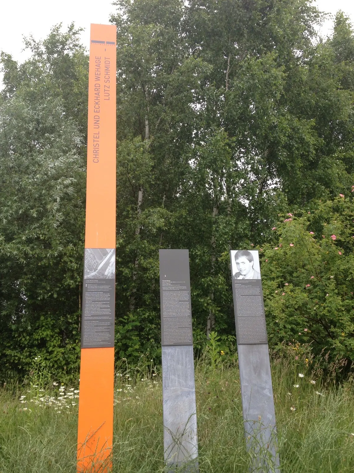 Tall orange column with the names Christel und Eckhard Wehage and Lutz Schmidt, beside grey posts with text panels, one with a portrait, in tall grass