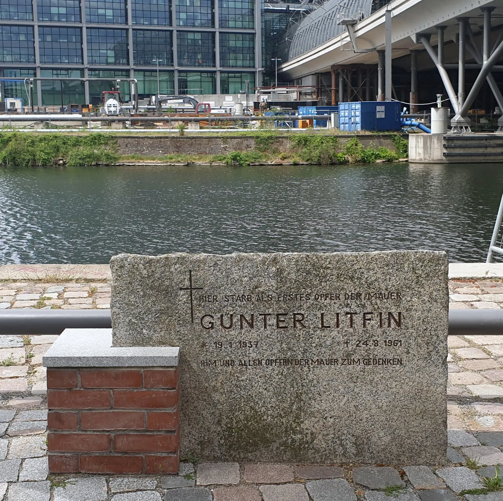 Granite stone with a small brick pedestal on a canal-side path, inscribed "Hier starb als erstes Opfer der Mauer Günter Litfin", with modern buildings across the water