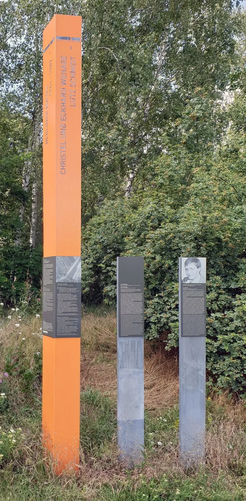 Tall orange column bearing the names Christel und Eckhard Wehage and Lutz Schmidt beside two dark steles, one with a portrait, in front of birch trees and bushes