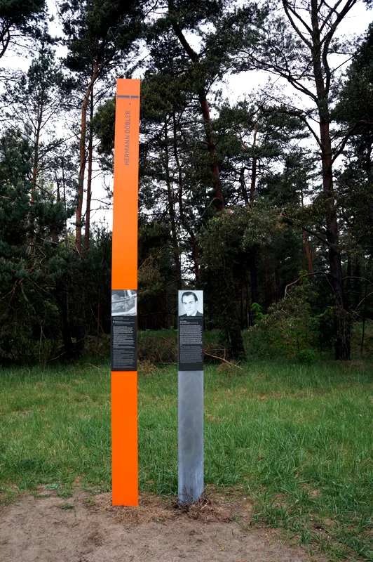 Tall orange column inscribed 'Hermann Döbler' beside a grey post with his portrait panel, on a grassy clearing in front of pine trees