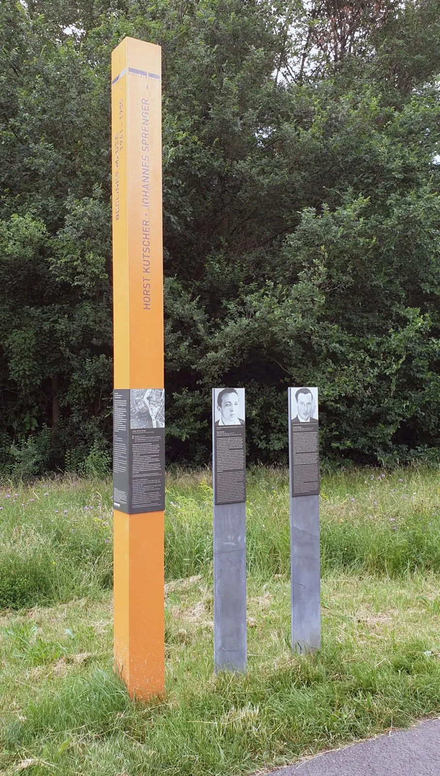 Tall orange column inscribed 'Horst Kutscher · Johannes Sprenger' beside two grey posts with portrait panels, in a meadow in front of trees