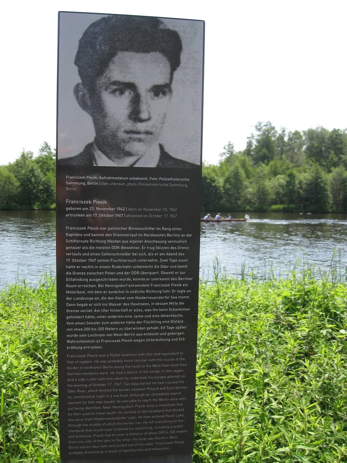 Dark panel with his portrait and text standing in tall grass on a riverbank, with a rowing boat on the water behind